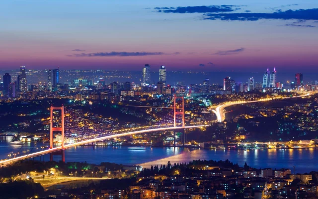 Wide-angle night view of the Bosphorus Bridge and the illuminated Istanbul skyline reflected on the water.